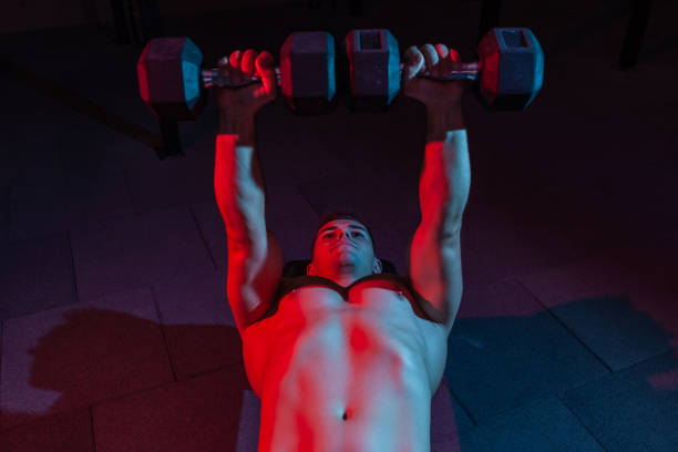 Muscular powerful man training bench press with dumbbells exercise in red blue neon light