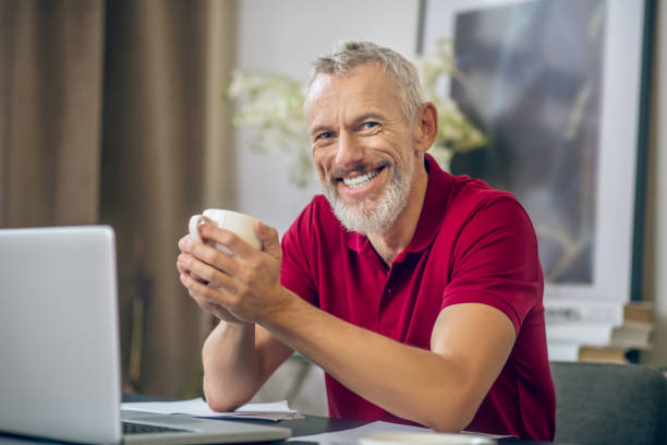 istock Grey-haired man sitting with a cup in hands and smiling 1290884362