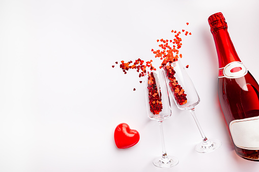 Two champagne glasses with splash of red heart shaped confetti over white background, top view with copy space. Valentine's Day concept