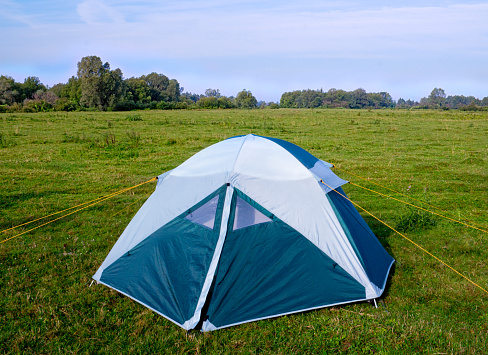 Practical single camping tent of blue-white color on the green meadow by the river.\