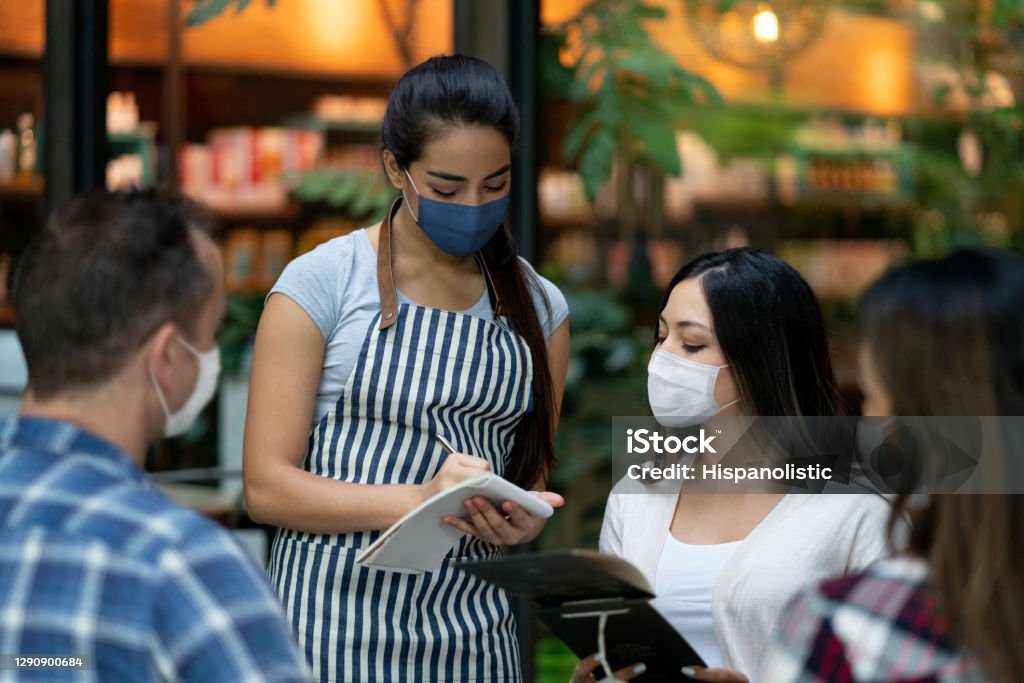 Waitress working at a restaurant during the pandemic and wearing a facemask Latin American waitress working at a restaurant during the COVID-19 pandemic and wearing a facemask while taking the order of the customers Restaurant Stock Photo