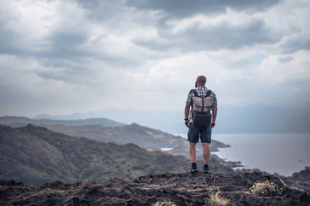 istock Man with backpack stands alone on the rocks at sunset 1290905401