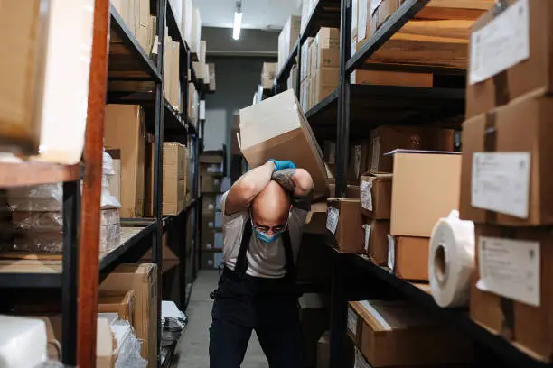 Endangered warehouse worker receiving a hit from the box falling off the shelf, protecting his back, shielding with both hands. Bent forward.