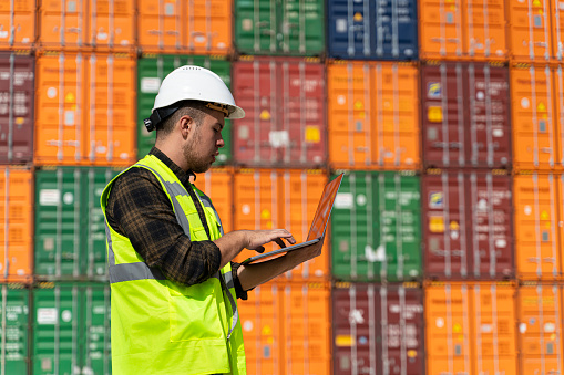Engineer man with white safety helmet and worker west checking cargo freights in front of colorful cargo container stacks in shipping port.