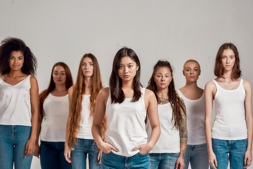 Portrait of beautiful young asian woman in white shirt looking at camera. Group of diverse women standing isolated over grey background. Diversity concept. Front view. Selective focus