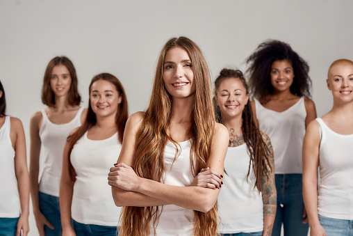 Portrait of beautiful young caucasian woman with long hair in white shirt smiling at camera. Group of diverse women posing, standing isolated over grey background. Diversity concept. Selective focus