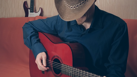 Man in cowboy hat and blue shirt plays a red acoustic guitar and sings. Electric guitar can be seen in background. Concept leisure and country music