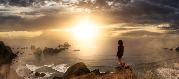 Ecola State Park, Cannon Beach, Oregon, United States. Adventurous Girl standing at the Rocky Cliff on Pacific Ocean Coast. Dramatic Colorful Sunset Sky.