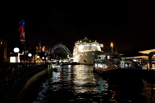 Sydney, New South Wales, Australia, April 19, 2017: The massive size of a Cruise Ship, Voyager of the Seas at Night with the Sydney Harbour Bridge in the Background. Docking at Circular Quay Cruise Ships are regular visitors to Sydney.