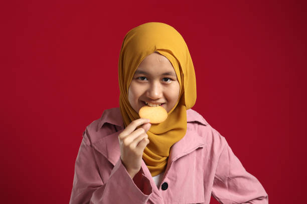 Young Asian muslim girl wearing hijab smiling at camera while eating biscuit cracker Young Asian muslim girl wearing hijab smiling at camera while eating biscuit cracker, against red background eating child cracker asia stock pictures, royalty-free photos & images