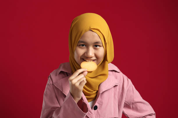 young asian muslim girl wearing hijab smiling at camera while eating biscuit cracker - eating child cracker asia imagens e fotografias de stock