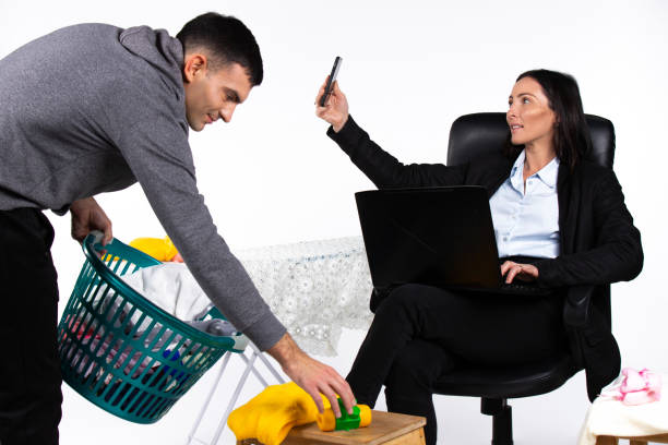 un uomo raccoglie cose in casa mentre la sua ragazza, un'imprenditrice, lavora a un laptop e si fa un selfie. rompere gli stereotipi di genere. - independence business women manual worker foto e immagini stock