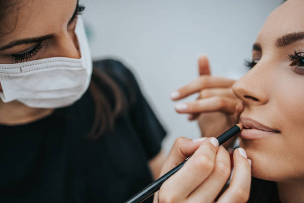 istock Make up artist with face protective mask applying professional make up of beautiful middle age brunette  woman. Coronavirus pandemic lifestyle. 1290972294