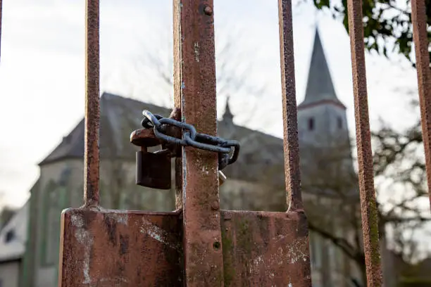 Photo of iron link chain with two locks lock a rusty iron gate, symbol for lockdown during covid-19 crisis, church in the background