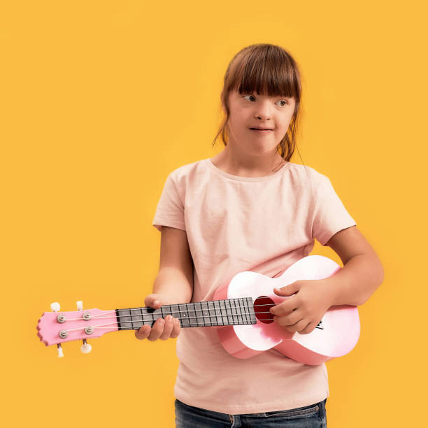 istock Cheerful disabled girl with Down syndrome looking aside while playing ukulele, standing isolated over yellow background 1290978157