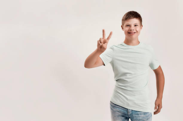 istock Happy disabled boy with Down syndrome smiling at camera, showing peace sign with one hand while standing isolated over white background 1290978868