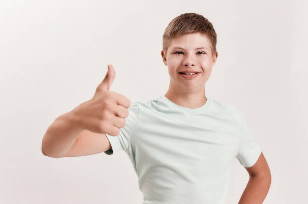 istock Joyful disabled boy with Down syndrome smiling at camera, showing thumbs up with one hand while standing isolated over white background 1290978970