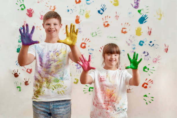 gosses heureux, garçon et fille handicapés avec le syndrome de down souriant à l’appareil-photo, affichant leurs mains peintes dans les peintures colorées pour des impressions de main sur le mur - child cheerful little girls down syndrome photos et images de collection