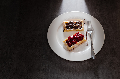 Tasty waffles with fruit and chocolate topping, white plate on dark background under strong, directional back light.