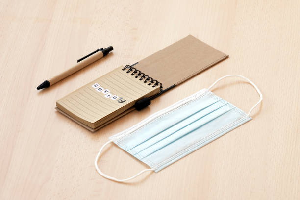 istock Flat lay with copy space. A face mask, a spiral notepad with “Covid-19” written on it and a pen are arranged on a wooden table. 1291016642