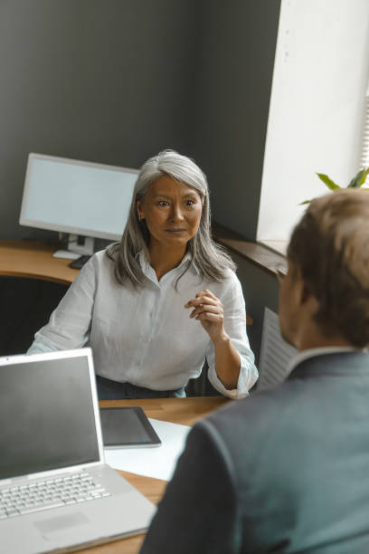 istock Two lead supervisors cheerfully talking together sitting one against another. Asian mature business woman talking to a manager working on computer with a black screen sitting at the modern office 1291017997