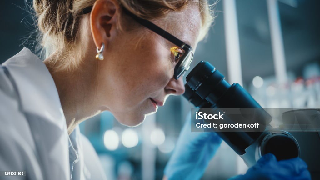 In Bright Medical Science Laboratory: Beautiful Microbiologist Wearing Glasses Looks Under Microscope Analyzing Sample. Brilliant Scientist, Doctor, working with High-Tech Equipment. Close-up Shot Scientist Stock Photo