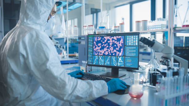 istock Medical Laboratory: Microbiology Scientist Wearing Sterile Coverall, Face Shield and Mask Works on Computer Developing Vaccine, Drugs and Antibiotics. Screen Shows High-Tech Concept for DNA research 1291031234