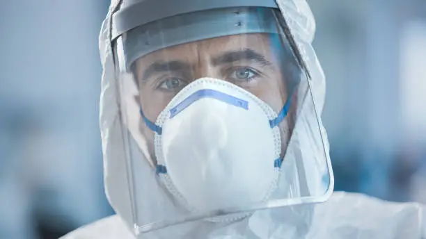 Photo of Dramatic Shot of Heroic and Overworked Medical Worker Wearing Coverall, Face Mask and Shield Looks Up at the Camera with His Piercing but Hopeful Eyes. Health Worker Fighting against Pandemic