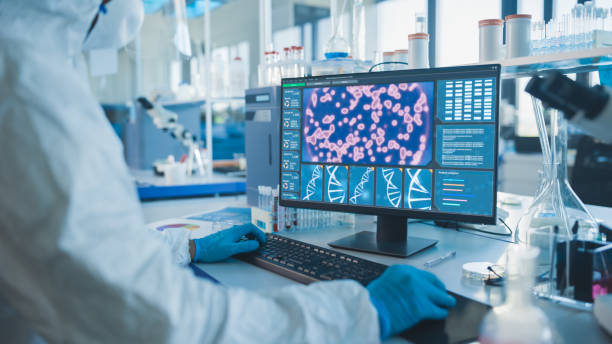 istock Medical Laboratory: Microbiology Scientist Wearing Sterile Coverall, Face Shield and Mask Works on Computer Developing Vaccine, Drugs and Antibiotics. Screen Shows Concept for DNA research. 1291031359