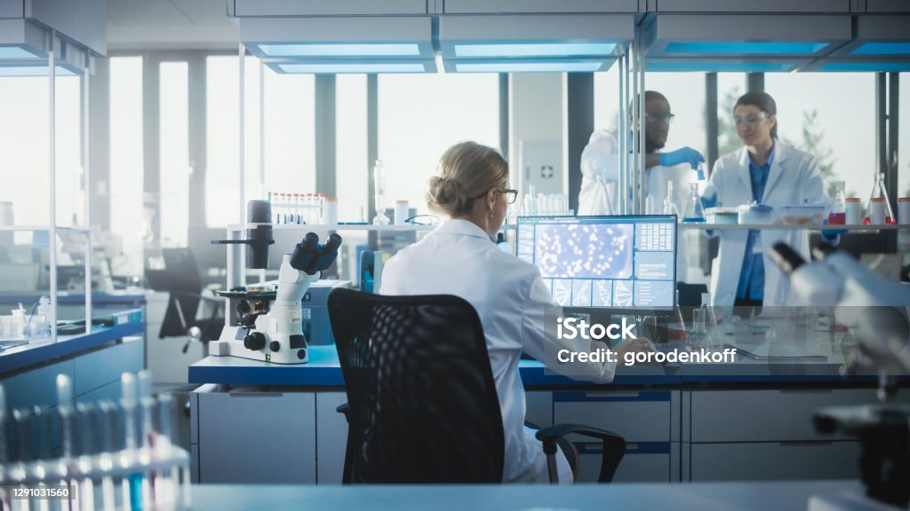 Medical Laboratory with Team of Scientists Working. Microbiologist is Using Personal Computer, Screen Shows High-Tech Concept for DNA research. High-Tech Biotechnology Research. Back View Shot Laboratory Stock Photo