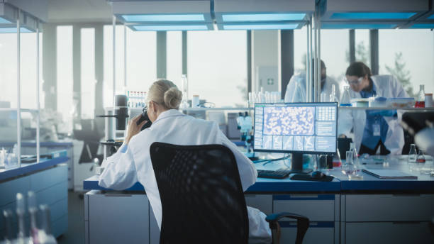 istock Medical Laboratory with Team of Scientists Working. Microbiologist Looking under Microscope and working on Computer. Developing Drugs, Gene Editing, High-Tech Biotechnology Research. Back View Shot 1291031590