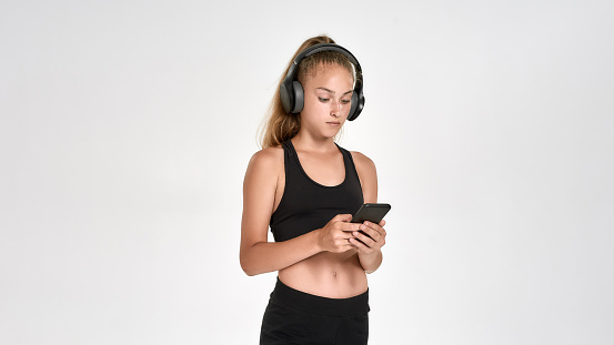 Portrait of cute sportive girl child wearing headphones, using smartphone while posing isolated over white background. Sport, training, fitness concept. Web Banner