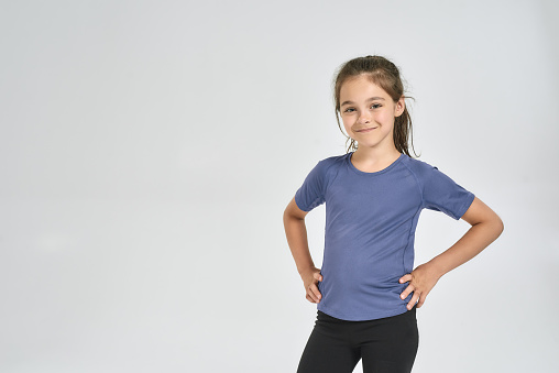 Little sportive girl child in sportswear smiling at camera, while standing isolated over white background. Sport, training, fitness, active lifestyle concept. Horizontal shot