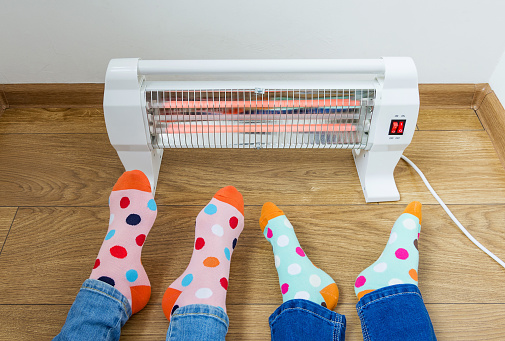 A young family wearing bright polka dot socks warms their cold feet near an electric heater. Infrared halogen heater at home.