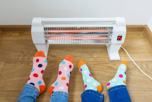 A young family wearing bright polka dot socks warms their cold feet near an electric heater. Infrared halogen heater at home. stock photo