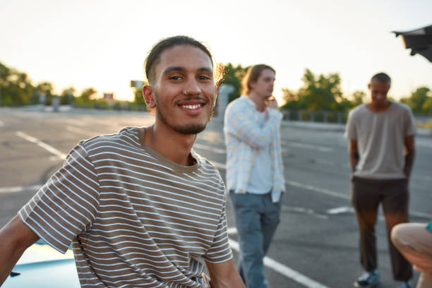 istock A close up of a casually dressed dark-skinned latina guy with a piercing smiling looking into a camera standing outside on a parking site with his friends on a background 1291048832