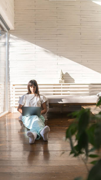 istock Young woman working on a laptop while sitting on a sunny window seat 1291052854