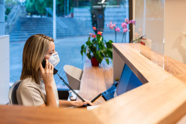 young woman answer the phone at the reception with coronavirus mask young woman answer the phone with coronavirus protection mask at the reception of modern fitness club or health centre medical office lobby stock pictures, royalty-free photos & images
