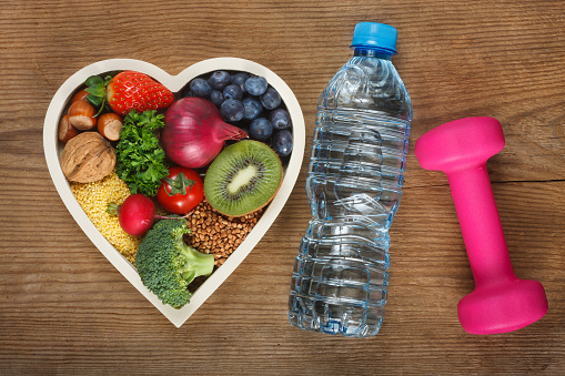 Healthy food in heart shaped bowl, bottle of water and dumbbell. Food such as blueberries, red onion, strawberry, parsley leaves, hazelnuts, walnut, tomato. kiwi, millet, buckwheat, radish, broccoli.