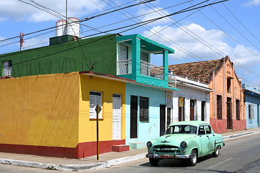 A row of colorful traditional houses with a paved street and Cuban vintage car against blue sky in Trinidad, Sancti Spiritus Province of Cuba