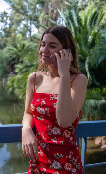 Beautiful young woman wears a red dress. She is talking on the phone in the park