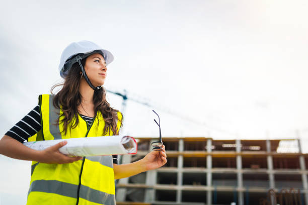 istock Young female builder/ engineer / architect / inspector / manager at construction site, holding a construction plan and looking at the sky. Architecture / construction industry concept. 1291070552