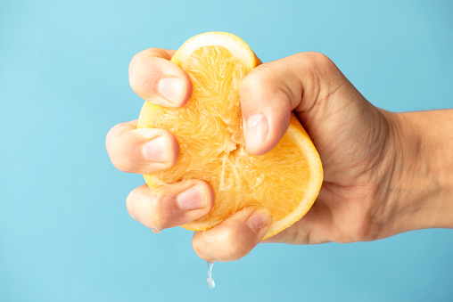 Close up of slice of  hand squeezing orange fruit in front of blue background.