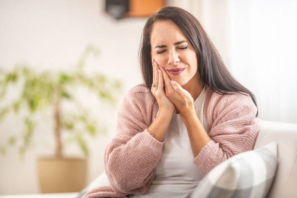 brunette s’assied à la maison avec un mal de dents retenant sa joue droite avec des mains. - rage de dents photos et images de collection