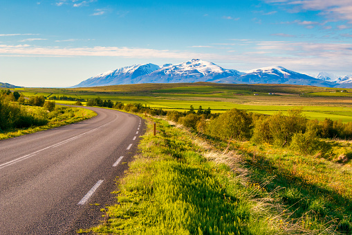 The bend of the asphalt road among green meadows in front of the mountain peaks on Iceland.