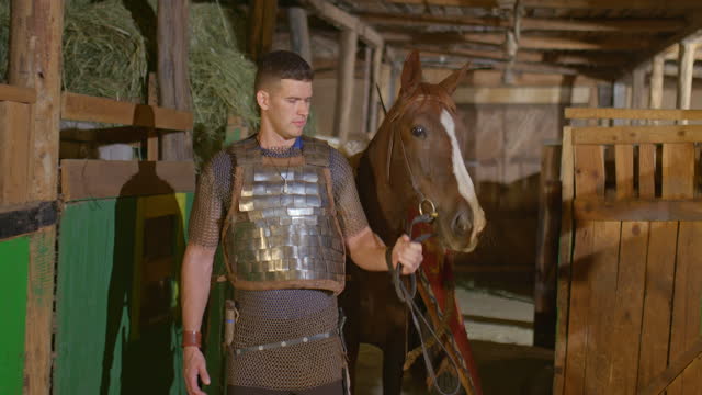 Portrait, a warrior in armor and chain mail leads the horse out of the stable. Shield and spears hang on a horse, a Russian warrior. 4k, ProRes