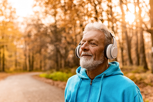 Senior man on a jogging pursuit standing on forest road with headsets on. Listening to music and relaxing with eyes closed.