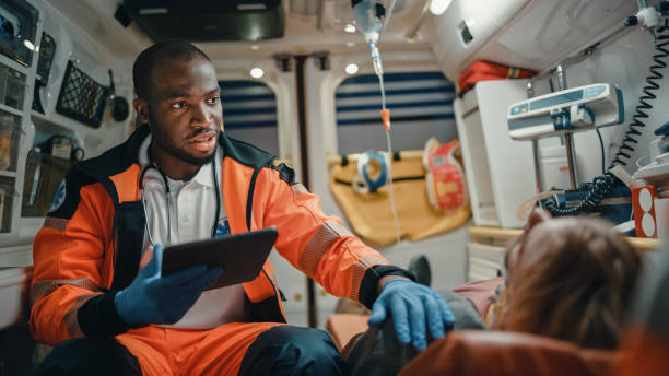 paramédic professionnel afro-américain noir d’ems utilisant l’ordinateur de tablette pour remplir un questionnaire pour le patient blessé sur le chemin de l’hôpital. l’assistant de soins médicaux d’urgence travaille dans une ambulance. - véhicule de service durgence photos et images de collection