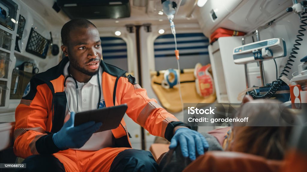 Black African American EMS Professional Paramedic Using Tablet Computer to Fill a Questionnaire for the Injured Patient on the Way to Hospital. Emergency Medical Care Assistant Works in an Ambulance. Paramedic Stock Photo