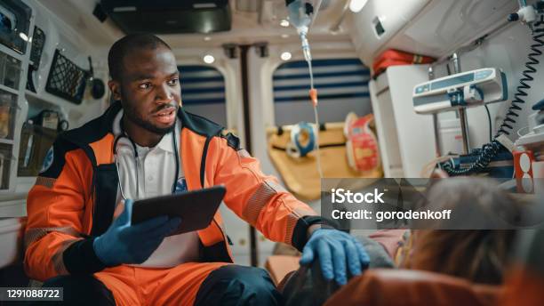 Black African American Ems Professional Paramedic Using Tablet Computer To Fill A Questionnaire For The Injured Patient On The Way To Hospital Emergency Medical Care Assistant Works In An Ambulance Stock Photo - Download Image Now